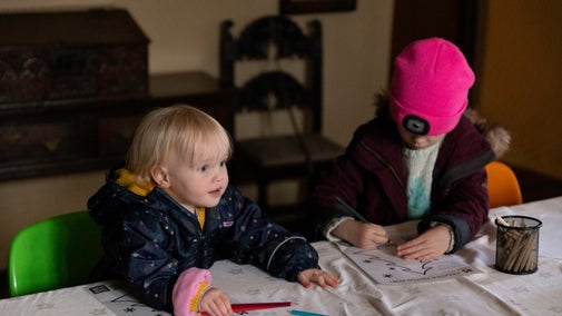 Two children sat at a table colouring. There is dark wooden furniture in the background.